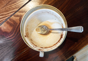An empty dirty cappuccino coffee cup, full of creamy coffee remnants , served on a wooden rustic brown coffee shop table. Organic fair trade coffee photographed from above top view