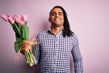 Young african american afro romantic man with dreadlocks holding bouquet of pink tulips with a happy face standing and smiling with a confident smile showing teeth