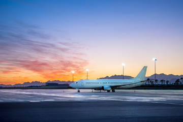 Fototapeta na wymiar Plane Parking Outside The Terminal In Airport In Resort Town, International Airlines . White Passenger Airplane Side View, Airport Lights, Sunset Over The Mountains.