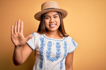 Young beautiful asian girl wearing casual t-shirt and hat standing over yellow background showing and pointing up with fingers number five while smiling confident and happy.