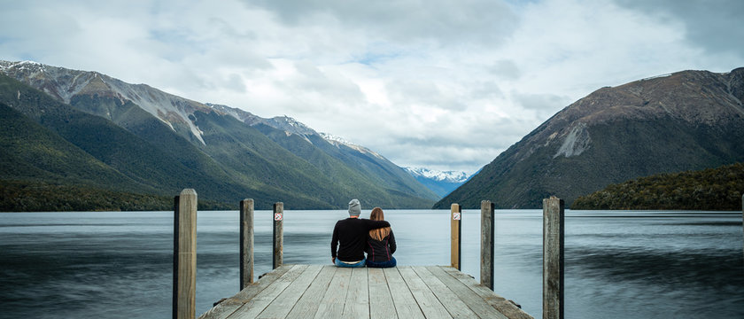 Rotoiti Lake, New Zealand, October 8, 2019: Beautiful panoramic picture of a loving couple embracing each other sitting at the end of a yetty with snowy mountains in the background