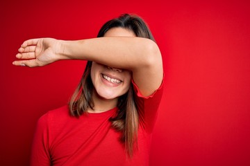 Obraz na płótnie Canvas Young beautiful brunette girl wearing casual t-shirt over isolated red background Smiling cheerful playing peek a boo with hands showing face. Surprised and exited