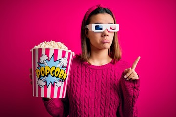 Young beautiful girl watching movie using 3d glasses eating box with popcorns Pointing up looking sad and upset, indicating direction with fingers, unhappy and depressed.