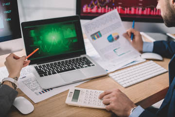 Cropped view of information security analysts using calculator and graphs on computer monitors while working in office