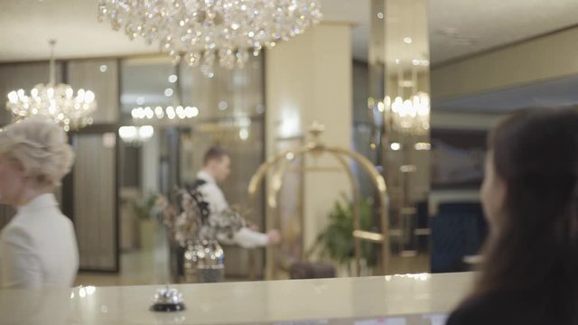 Back view of unrecognizable receptionist welcoming wealthy clients in luxurious hotel and giving them room key card. Happy man and woman smiling and leaving as bellboy passing at the background.