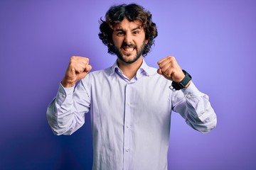 Young handsome business man with beard wearing shirt standing over purple background angry and mad raising fists frustrated and furious while shouting with anger. Rage and aggressive concept.