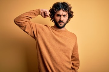 Young handsome man with beard wearing casual sweater standing over yellow background Strong person showing arm muscle, confident and proud of power