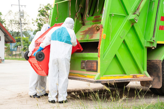 Two Garbage Men In Hazmat PPE Protective Clothing Wear Medical Rubber Gloves Working Together On Emptying Dustbins For Trash Removal With Truck Loading Waste And Trash Bin,Coronavirus Disease 2019.