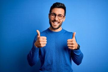 Young handsome man with beard wearing casual sweater and glasses over blue background success sign...