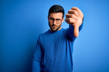Young handsome man with beard wearing casual sweater and glasses over blue background looking...