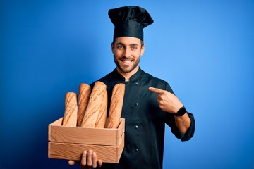 Young cooker man with beard wearing uniform holding box with bread over blue background with surprise face pointing finger to himself