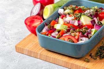 Raw sliced vegetables in a baking dish on the kitchen table. Zucchini, tomatoes, onions, capers and herbs are prepared for baking in the oven. Tasty and healthy vegan and vegetarian food.