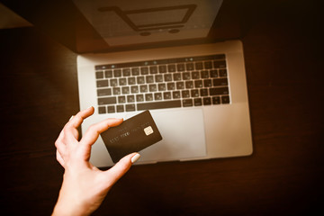 woman's hand is holding a black credit card above silver laptop standing on wooden table