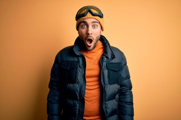 Young handsome skier man with beard wearing snow sportswear and ski goggles afraid and shocked with surprise and amazed expression, fear and excited face.