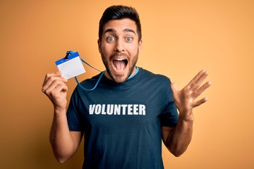 Handsome volunteer man with beard holding id card identification over yellow background very happy and excited, winner expression celebrating victory screaming with big smile and raised hands