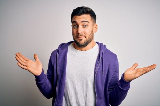 Young handsome man wearing purple sweatshirt standing over isolated white background clueless and confused expression with arms and hands raised. Doubt concept.
