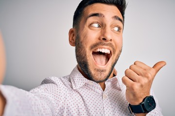 Young handsome man wearing shirt making selfie by the camera over white background pointing and showing with thumb up to the side with happy face smiling