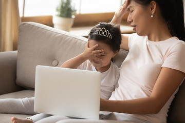 Asian little daughter and mother sit on couch in living room watching cartoon on laptop closed eyes...