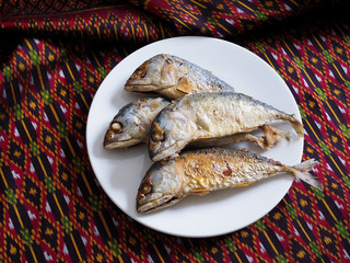 Top view of Thai food with fried mackerel fish or rastrelliger brachysoma in white plate on traditional Thai fabric background.
