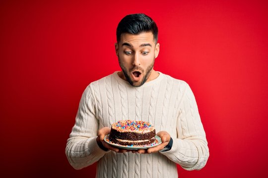 Young handsome man holding colorful birthday sweet cake over red isolated background scared in shock with a surprise face, afraid and excited with fear expression