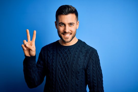 Young handsome man wearing casual sweater standing over isolated blue background smiling looking to the camera showing fingers doing victory sign. Number two.