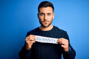 Young handsome man holding paper with solidarity message over blue background with a confident...