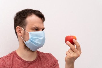 Caucasian man in facial surgical medical protective mask thinks how to bite an apple in a mask.