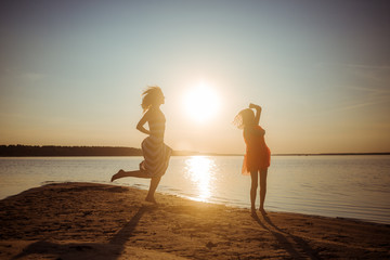 Happy mom and daughter in dresses are jumping and dancing on the beach during sunset. Good...