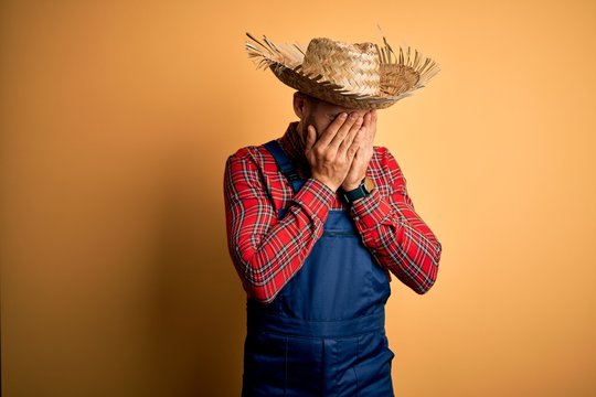 Young rural farmer man wearing bib overall and countryside hat over yellow background with sad expression covering face with hands while crying. Depression concept.