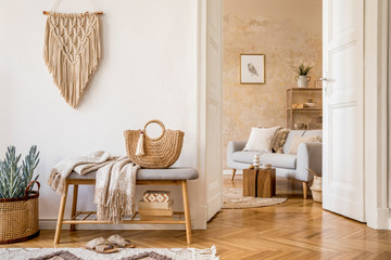 Scandinavian interior of open space with wooden bench, grey sofa, pillows, palid, picture frame, macrame, plant, books, carpet, decoration and elegant personal accessoreis in stylish home decor.