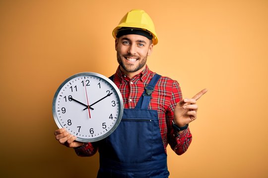 Young builder man wearing safety helmet holding big clock over yellow background very happy pointing with hand and finger to the side