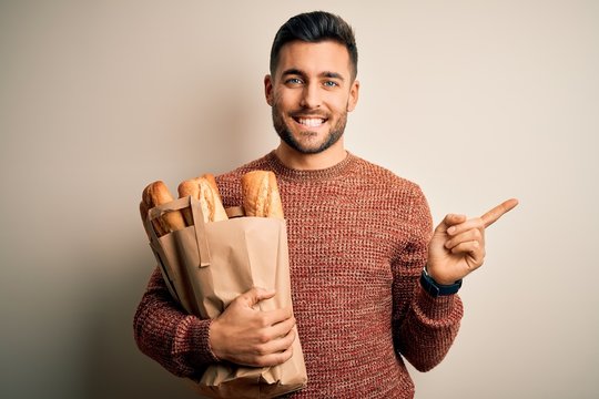 Young handsome man holding groceries paper bag of fresh baguette bread over isolated background very happy pointing with hand and finger to the side