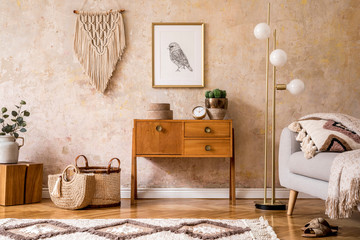 Modern retro composition of living room with wooden vintage commode, grey sofa, gold lamp, macrame, carpet, pillows, gold mock up poster frame, plants, decoration and personal accessories.