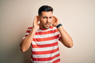 Fototapeta premium Young handsome man wearing casual striped t-shirt standing over isolated white background Trying to hear both hands on ear gesture, curious for gossip. Hearing problem, deaf