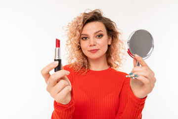 blond curly girl holds out lipstick and a mirror on a white background