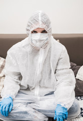 Person in protective white translucent suit, blue rubber gloves, medical mask sits at table at home and works or studies on laptop during quarantine. Remote work during coronavirus pandemic.