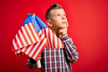 Young little american patriotic caucasian kid holding flag of USA over red background serious face...
