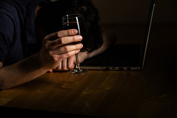 Depressed man on home office (in front of computer) holding glass of red wine. Working at home and alcoholic problem