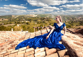 Junge Frau in blauen Kleid sitzt auf einem Dach und blickt in die italienische Landschaft bei...