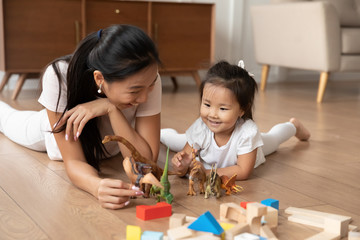 Lying on warm wooden floor asian mom play game with little daughter holds dinosaurs toys heap of...