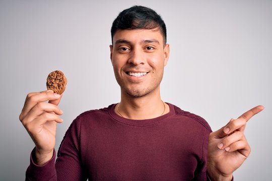 Young hispanic man eating chocolate chips cookie over isolated background very happy pointing with hand and finger to the side