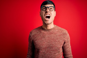 Young handsome hispanic man wearing nerd glasses over red background angry and mad screaming...
