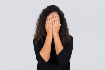 Curly haired brunette woman standing dressed in black  covering her face with her hands over gray...