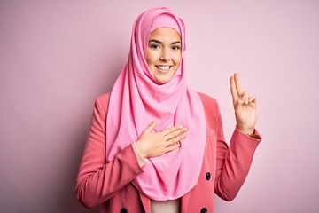 Young beautiful girl wearing muslim hijab standing over isolated pink background smiling swearing with hand on chest and fingers up, making a loyalty promise oath