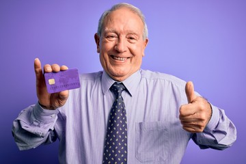 Senior grey haired business man holding credit card over purple background happy with big smile doing ok sign, thumb up with fingers, excellent sign