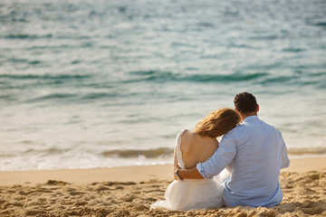 Newlyweds sitting close together at sunset on the beach in the sand, gazing out to sea. Bride and...