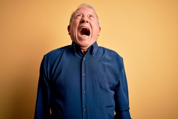 Grey haired senior man wearing casual blue shirt standing over yellow background angry and mad screaming frustrated and furious, shouting with anger. Rage and aggressive concept.