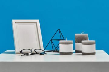 White coffee table with empty photo frame, three candles, decorative iron triangle and black rimmed glasses. Blue background. Close up, copy space