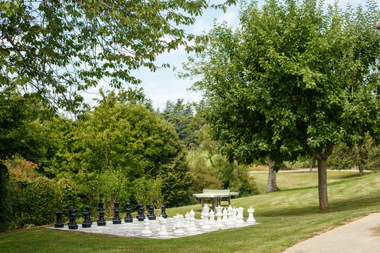 Giant Chess Figures Standing In Park. Chessboard Among Green Grass And Trees. Concept Of Intellectual Open Air Games, Leisure In Garden. Elegant Exterior Design