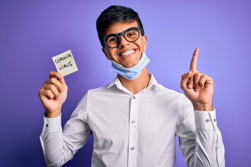 Young handsome man wearing medical mask holding reminder with corona virus message surprised with an idea or question pointing finger with happy face, number one